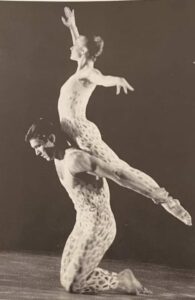 Black and white photo of Peter on his knees, lifts another dancer on his back. They are wearing unitards.