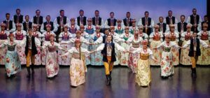 Large ensemble in traditional Polish attire, open their arms wide on a stage and step forward.