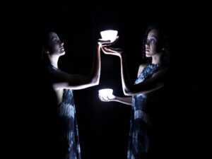Two dancers in a dark space and light blue dresses hold up 2 small lamps, one above the other