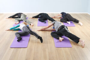 Five people on lilac yoga mats lay on their right side, extend their arms in front of their chest with hands together, bend right knee towards chest and extend left leg behind them.