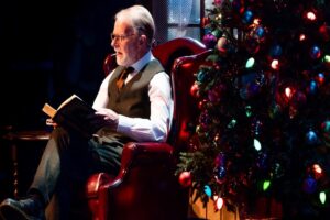 Senior performer sits on a red arm chair by a Christmas tree and reads a book.