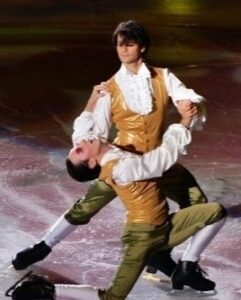 Two male-presenting ice-skaters dance together.