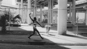 Black and white photo of a dancer in a calm street's sidewalk taking an elegant step forward while reaching one arm up behind them and one arm long in front of them.