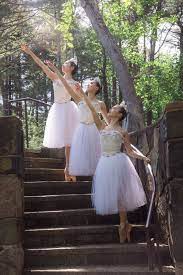 Three dancers on pointe in sous-sous on different levels of a staircase outdoors, wear white long tutus and look up over their right arms reaching on a high diagonal.