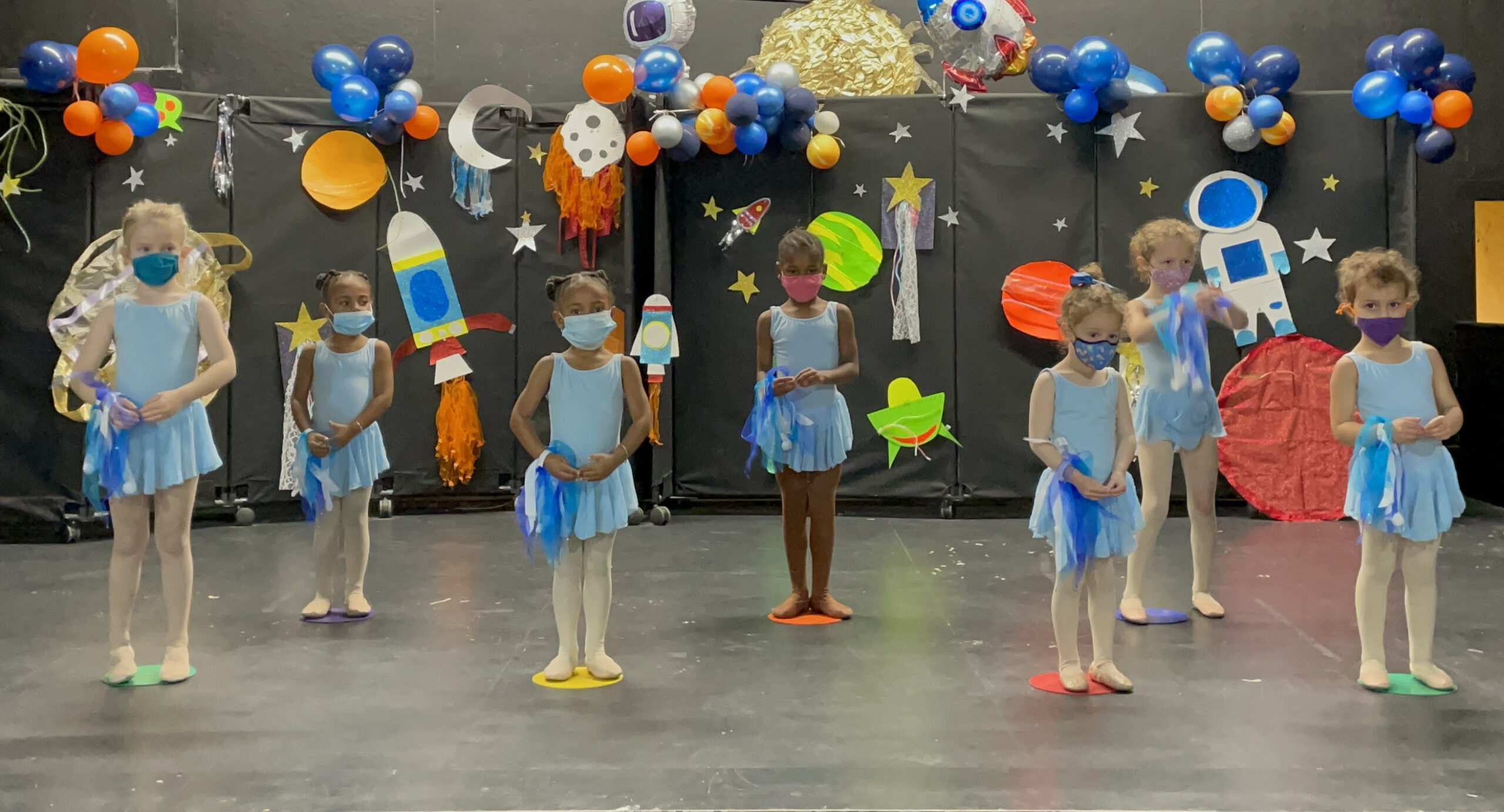 Studio decorated with an outer space theme and group of young dancers in blue leotards and skirts stand on colorful spots.
