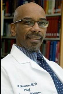 Dr. Shannon, a bald Black man with a goatee wearing glasses and a white physician's coat embroidered with his name, sits in front of a shelf of books.