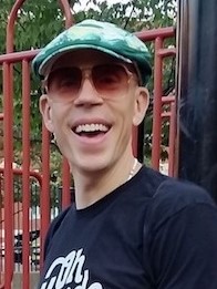 Aaron wears a hat and sunglasses and smiles at the camera.