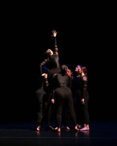 Nozama Dance Collective dancers in all black stand and hold up one of the dancers who reaches right arm up.