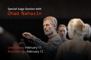 Ohad Naharin looks towards his left hand and reaches his left arm towards the left side of the photo