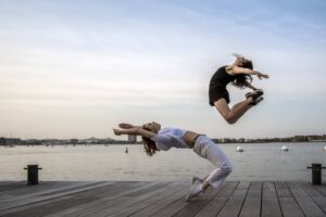Two dancers by a waterfront on a pier: one in a black romper jumps and reaches arms and legs back, arching her back as the other, wearing a white shirt and pants hinges back over the tops of her feet and reaches arms over head.
