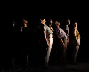 Dark photo of BDT company members in earth tones wearing face masks and standing, slightly hinged back, facing a side light on stage left.