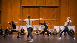 Dancers in a large studio space lunge forward and open chests in a circular shape while others sit and observe.