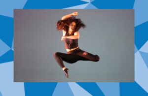 Dancer in black leggings and black cropped top jumps with both legs bent and staggered with one arm bend over head and the other bent in front of chest with long hair swishing to the sides.