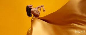 Alvin Ailey dancer in a long yellow dress looking up and bending arms in front of chest in a gentle gesture.