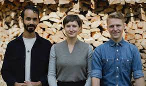 Jake, Laurel and Nic photographed from waistline up in front of a large pile of fire wood.