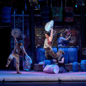 Performance photo of STOMP with one performe jumping with legs bent back and arms up holding on to garbage tin lids. Another performer also holds on to tin lids, opens arms wide and stands on one leg as the other is bent and lifted.
