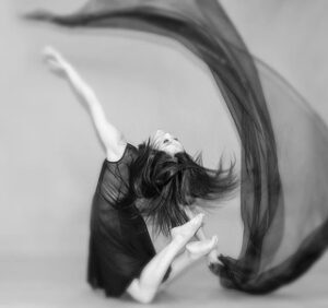 dancer with long straight hair on knees, brings toes towards head as they arch back and toss a long lofty fabric back.