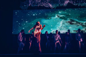 Dancers on a dim-lit stage and a galactic background projected. One of the dancers jumps with one leg bent and the other straight down.