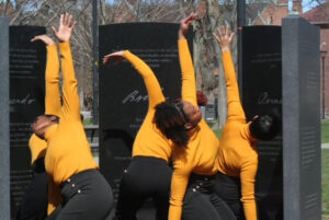Dancers in yellow turtlenecks and black pants reach one arm up and slightly hinge sideways.