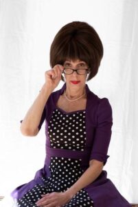 Lynn Modell wearing a polka-dot dress sits and hols on to glasses with one hand just underneath the eye-line.