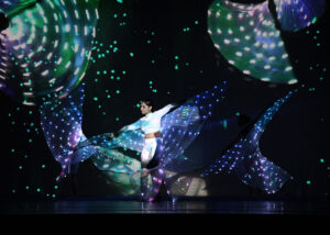 Photo of performer in white costume dancing with lights strung together on a long fabric and sticks.