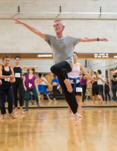 Daniel in class with other dancers jumps with one leg bent and arms extended to the sides.