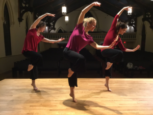 Three dancers on a wooden floor stage in black capri length pants and red shirts hold one leg bent in a parallel passé while on relevé and around their arms above their head as they curve their upper body on a diagonal towards their left side.