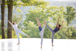 Three dancers lift their legs and arms joyfully on the Jacob's Pillow outdoor stage with trees in the background.