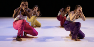 Four dancers in different colored pants crouched down with hands over one eye was if looking through a telescope.