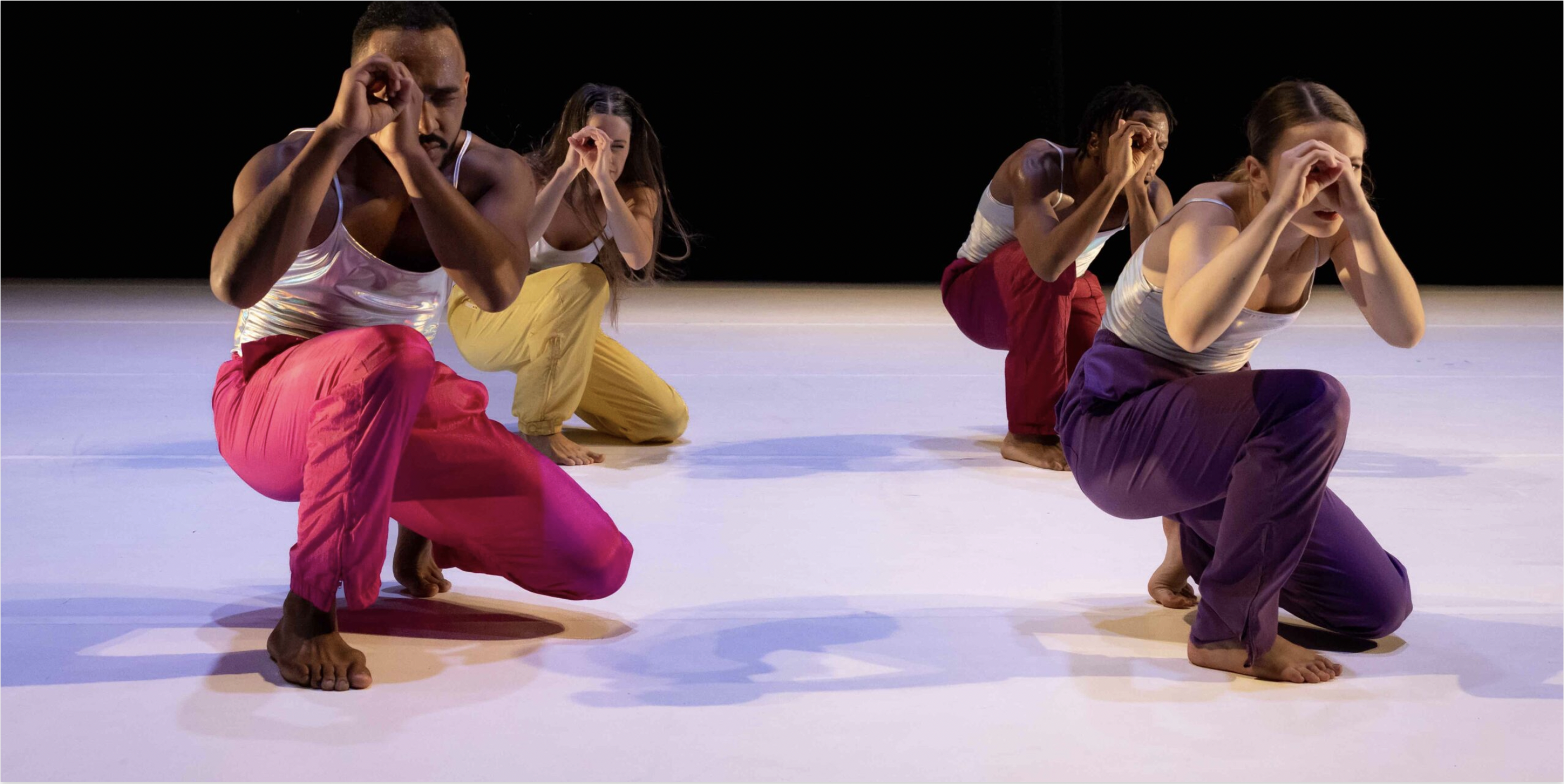 Four dancers in different colored pants crouched down with hands over one eye was if looking through a telescope.
