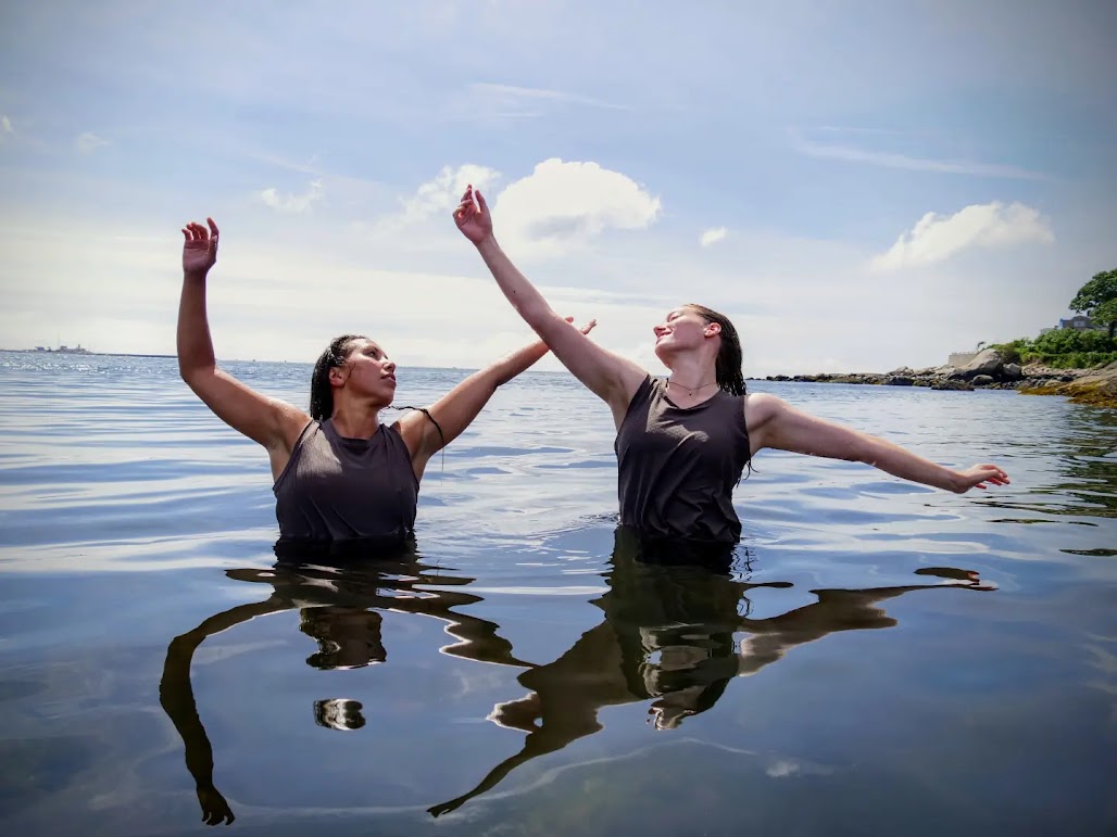 Two dancers immersed in a body of water reaching arms up.