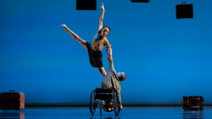 Dancer in a wheelchair reaches one arm up holding on to another dancer who leaps with both legs straight in a straddle.