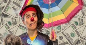 Clown with red nose, headband and floppy brown hair grimaces under a rainbow umbrella and background of paper money.