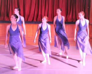 Annex Contemporary Dance company in leotards and long loose skirts on stage looking over right shoulder while taking a step on left foot.