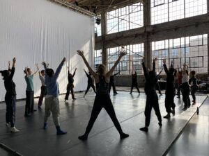 Many dancers silhouetted by natural light in a large circle stand with legs wide and reach arms up.