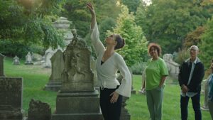 Dancer in white long sleeve top reaches one arm up gently and looks up at fingers, as two people observe in the background of the cemetery.