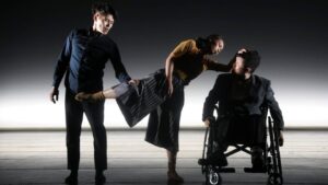 Three dancers on a dim-lit stage, from left to right: one dancer stands and holds the second dancer's leg; the second dancer lifts one leg and leans over towards third dancer in a wheelchair.