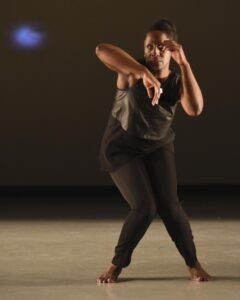 Deborah Goffe in all black on stage bending knees towards each other and bending arms to form parallel diagonal lines.