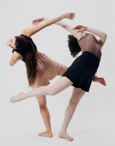 Two dancers in shorts and tank tops are back to back arching over left shoulder while lifting right leg bent behind them.