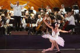 Ballerina in white swan costume kneels with arms open and bows. Orchestra in the background.