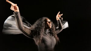 Black woman in dark clothes with fabric that looks like wings holds her arms up at an angle as if flying