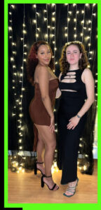 Two femme presenting latin ballroom dance instructors posing in front of a black background with string lights.