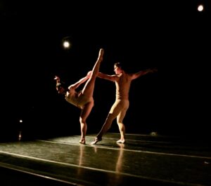 Dancers in pas de deux, one lifts straight leg up and the other holds the first's hand for support. Both dancers in tight fitted costumes on a dim-lit stage.