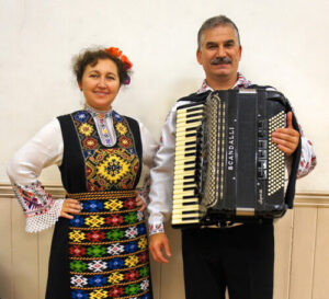 Iliana Bozhanova and Todor Yankova, holding an accordion, in traditional Bulgarian clothes stand side by side and smile.