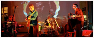Musicians in a band play together under a red-lit projection.