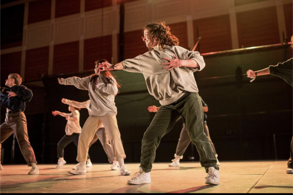 Dancers on a stage in baggy sweats reaching one arm out to their rights and left hand to heart
