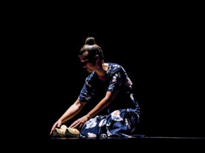 Photo of Laura Sanches in a dim lit stage kneeling and touching clogs