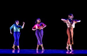 Three dancers in collared shirts and cropped pants of various colors cross the stage on relevée in a diagonal line against a black background.
