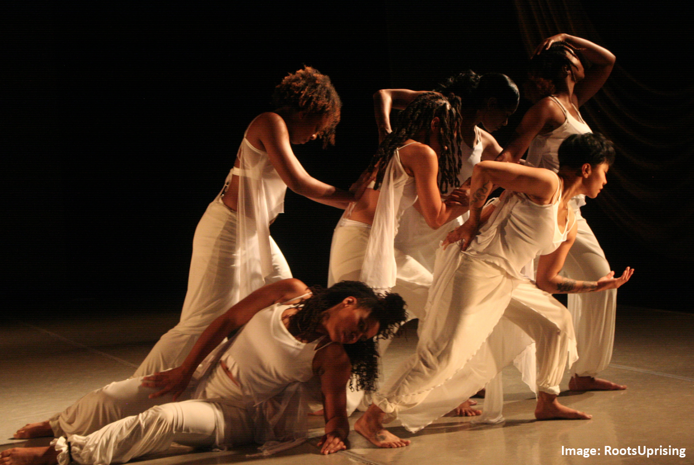 Group of dancers on stage in all white make shapes with their bodies and look towards stage left.