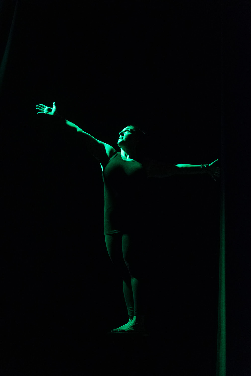 Sara Juli under green light reaches arms out and twists upper body.